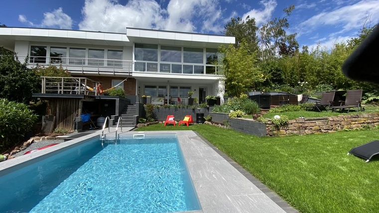 Ein modernes Haus mit großen Fenstern und einem gepflegten Garten. Im Vordergrund befindet sich ein Schwimmingpool, umgeben von Rasen und Liegestühlen.