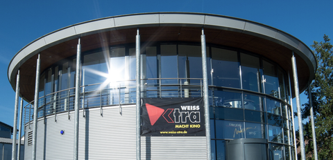 Ein modernes Gebäude mit großen Fenstern und einem geschwungenen Dach. An der Fassade hängt ein Banner mit dem Schriftzug "WEISS xtra macht Kino".
