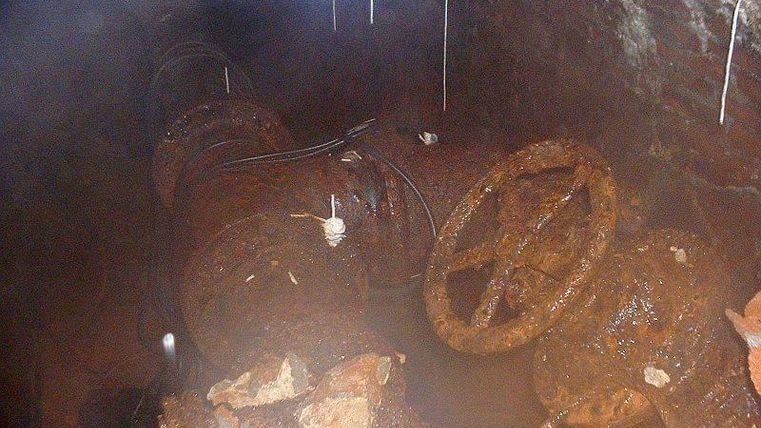Une canalisation rouillée enfouie dans le sol, surmontée d'un dispositif de commande de vanne. L'endroit semble humide et est entouré de briques effritées.