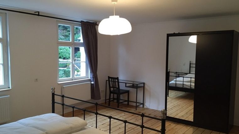 Une chambre simple avec un lit double, un bureau et un grand miroir. Des murs clairs et un sol en bois créent une atmosphère chaleureuse.