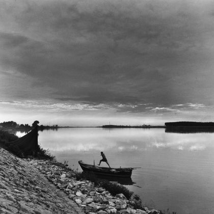 Ein stiller Fluss bei Sonnenuntergang, mit einem Boot am Ufer. Eine Person steht am Rand und schaut auf die spiegelnde Wasseroberfläche.
