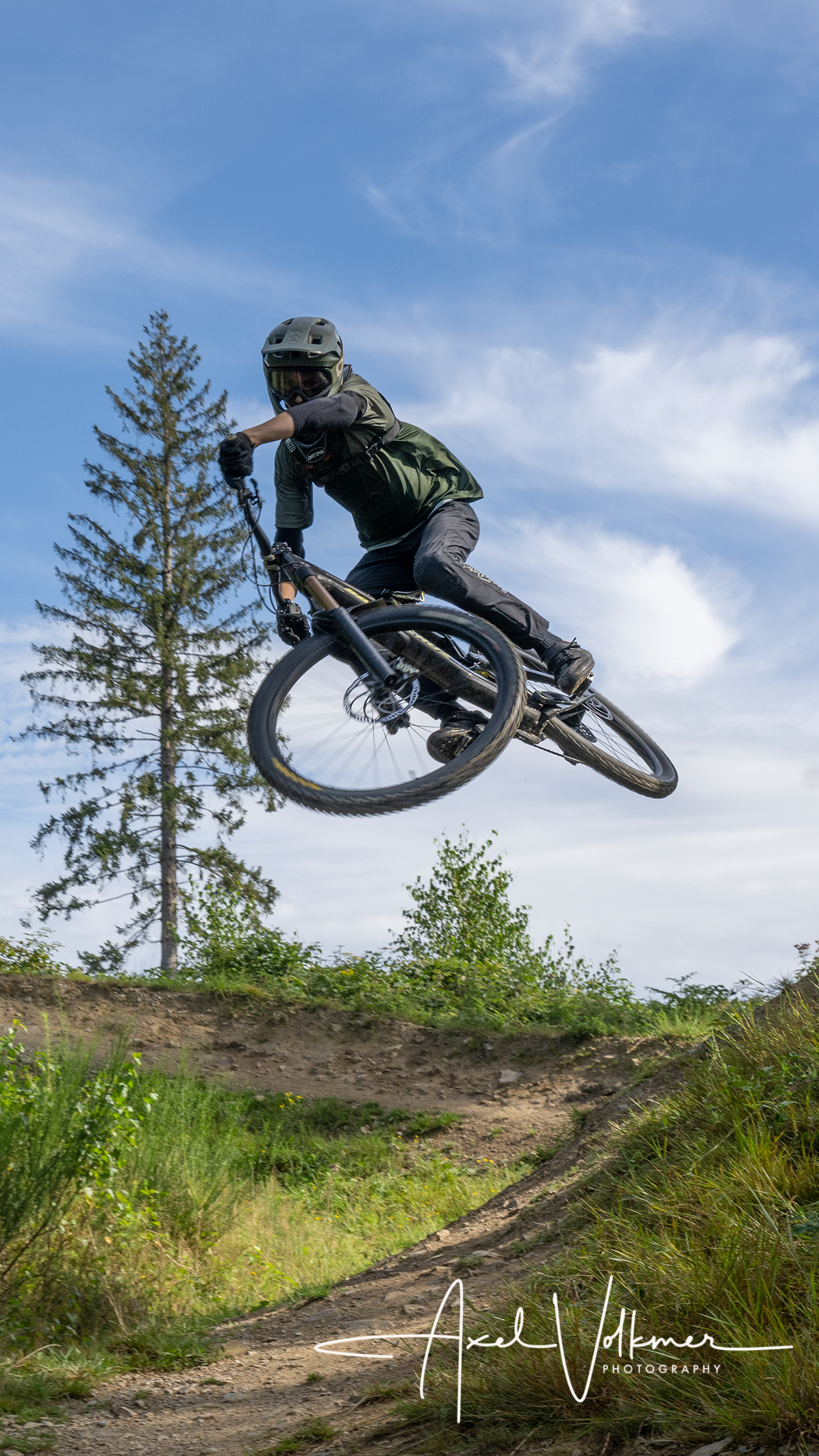 A mountain biker jumps over a hill with his bike. In the background, there are trees and a blue sky.