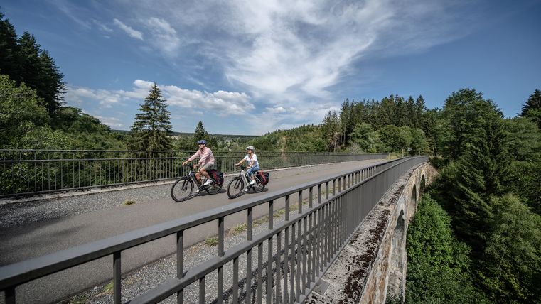 Zwei Radfahrer auf einem Viadukt in einer grünen Waldlandschaft.