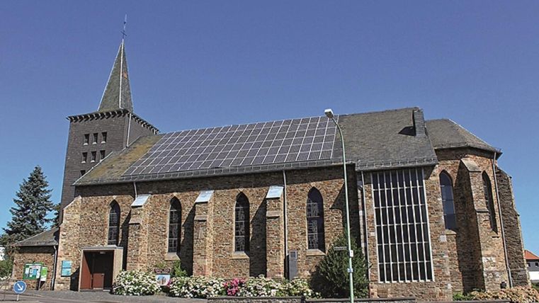 Eine Kirche mit einer steilen Spitze und Solarpanelen auf dem Dach. Die Fassade ist aus Stein und die Fenster sind in gotischem Stil gestaltet.