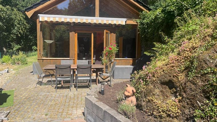 Een gezellig houten huis met grote ramen en een terras. De tuin is groen en goed onderhouden, omringd door planten en een stenen pad.