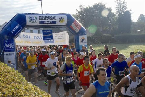 Eine große Gruppe von Läufern startet beim Monschau Marathon unter einem blauen Startbogen. Viele Teilnehmer tragen farbige Sportbekleidung und sind bereit für das Rennen.