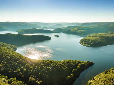 Eine malerische Landschaft mit einem ruhigen See und sanften Hügeln. Die Bäume reflektieren das Licht der Sonne und schaffen eine friedliche Atmosphäre.