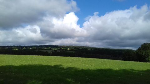 Eine weite grüne Wiese unter einem Himmel mit weißen Wolken und blauen Abschnitten. Im Hintergrund sind sanfte Hügel zu sehen.