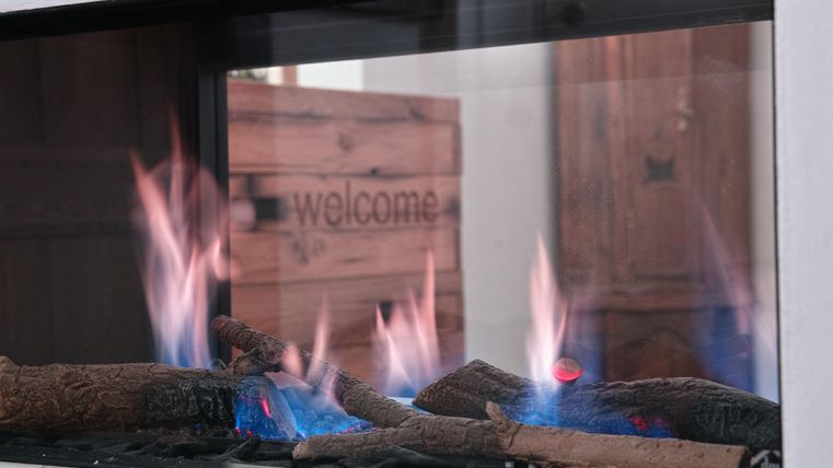 Ein moderner Kamin mit brennenden Holzscheiten. Im Hintergrund ist eine Holzkiste mit der Aufschrift "welcome" zu sehen.