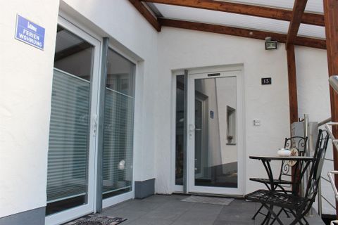 Ein einladender Eingangsbereich eines Ferienhauses mit gläserner Tür und Holzüberdachung. An einem kleinen Tisch stehen zwei Stühle, ideal für eine entspannte Auszeit.