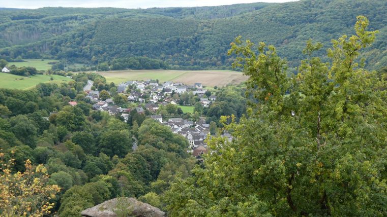 Eine malerische Landschaft mit sanften Hügeln und einem kleinen Dorf im Tal. Üppige Bäume umgeben die Szene und sorgen für eine ruhige Atmosphäre.
