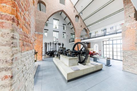 Ein historischer Raum mit Backsteinmauern und hohen Decken. In der Mitte steht eine große Maschine auf einem Betonblock.
