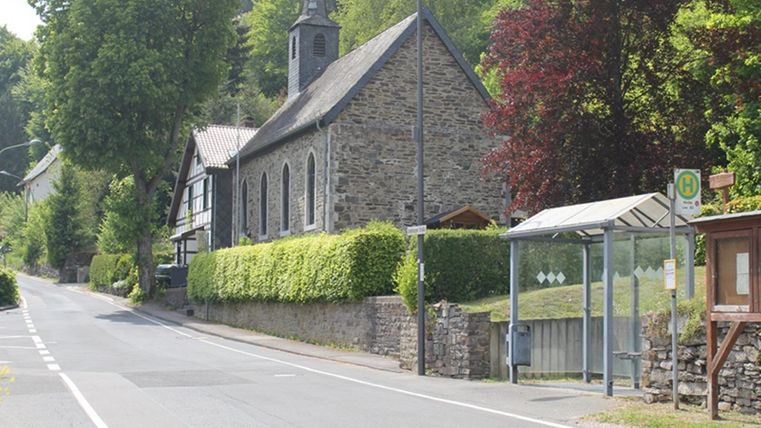 Eine ruhige Straße mit einer steinernen Kirche und grünen Bäumen. An der Seite befindet sich eine Bushaltestelle.