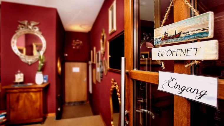 Ein stilvoller Eingangsbereich mit einer Holztür und einem Schild, das „Geöffnet“ anzeigt. An den Wänden hängen Bilder und es gibt einen großen Spiegel.