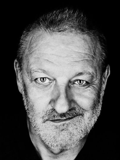 Ein Porträt eines lächelnden Mannes mit grauem Bart und kurzen Haaren. Der Hintergrund ist schwarz, was den Fokus auf sein Gesicht legt.