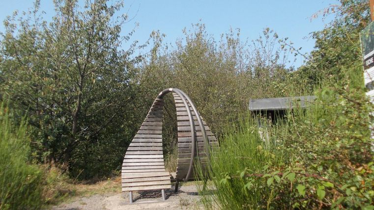 A wooden sculpture in the shape of an arch stands in a green environment with shrubs and trees. The sky is blue and clear.