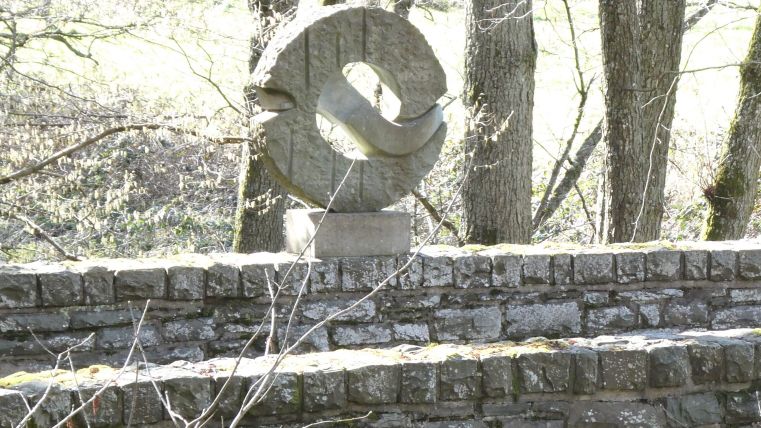 Eine moderne Steinskulptur auf einer Mauer, umgeben von Bäumen. Der Hintergrund zeigt eine ruhige, natürliche Umgebung.