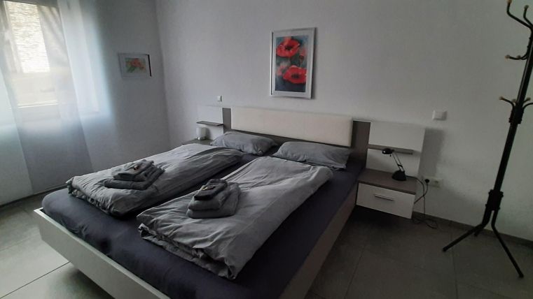 Ein modernes Schlafzimmer mit einem großen Bett und frischen Bettwaren. An der Wand hängt ein Bild mit roten Blumen und ein Kleiderständer steht im Raum.