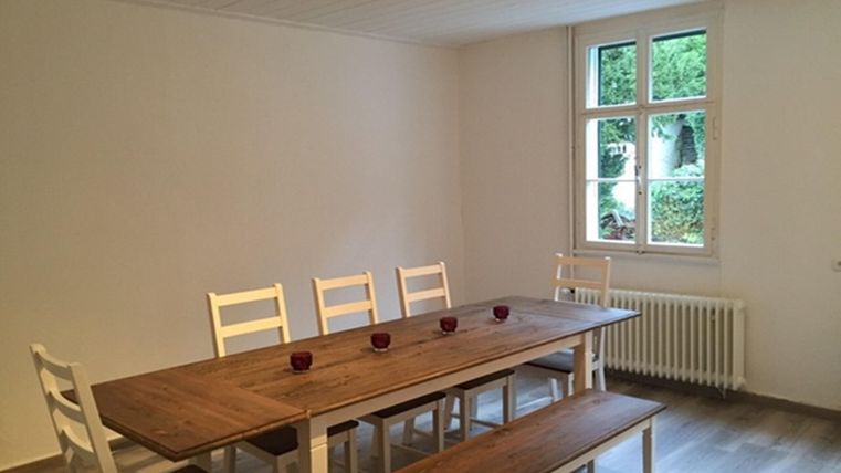 Une salle à manger lumineuse avec un plateau en bois et des chaises blanches. On peut voir une vue verte à travers la fenêtre.