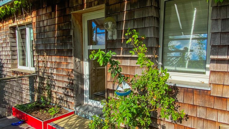 Ein Holzverschaltes Haus mit einem Eingang und zwei Fenstern. Vor dem Haus stehen rote Pflanzkästen mit grünen Pflanzen.