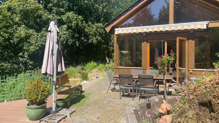 Een gezellig huis met grote ramen en een terras. De tuin is groen met planten en een tafel met stoelen.