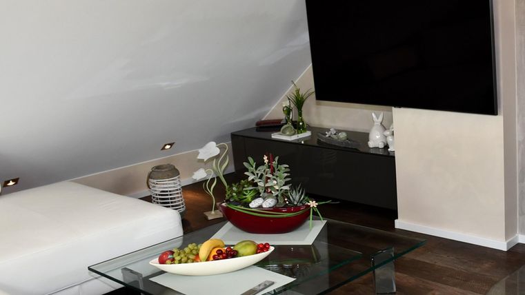 Un salon moderne avec une grande télévision et une table en verre. Sur la table se trouve un saladier de fruits et quelques plantes.