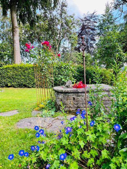 Ein schöner Garten mit bunten Blumen und einem alten Brunnen. Im Hintergrund stehen hohe Bäume und grüne Sträucher.