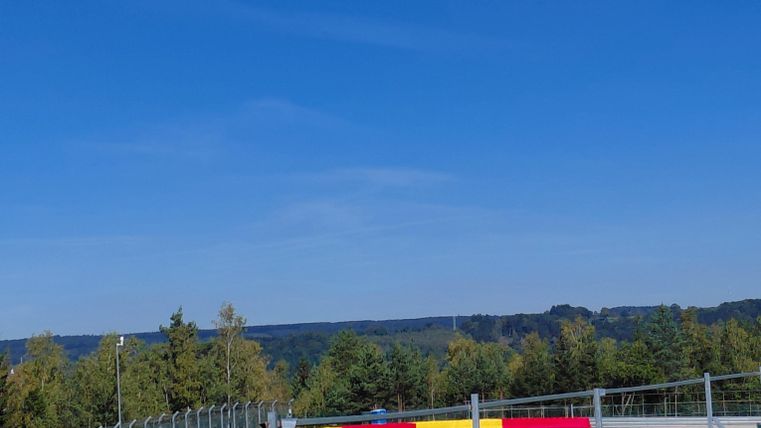 Eine Rennstrecke mit einem klaren blauen Himmel. Im Hintergrund sind Bäume und die umliegende Landschaft zu sehen.