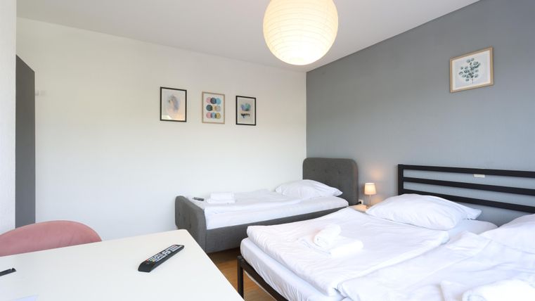 Ein einfaches Zimmer mit zwei Betten, einem Tisch und einem Stuhl. An der Wand hängen einige Bilder und eine Lampe sorgt für gemütliches Licht.