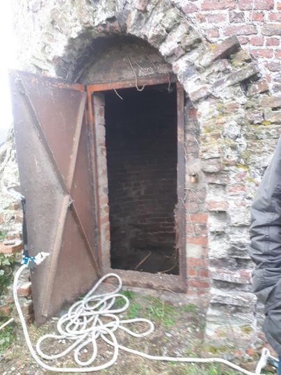 Eine alte, geöffnete Tür aus Metall führt in einen dunklen Raum. Vor der Tür liegen ein paar Schläuche auf dem Boden.