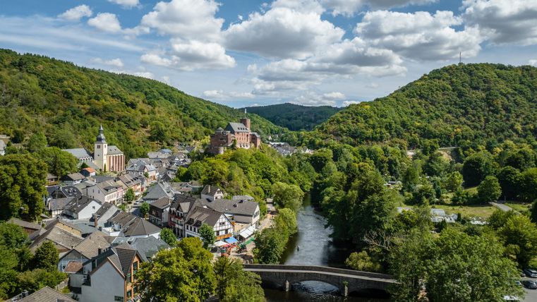 Une ville pittoresque au bord de la rivière, entourée de collines douces et de forêts denses. Les nuages glissent à travers un ciel clair, tandis que l'architecture de la ville incarne une histoire visible.