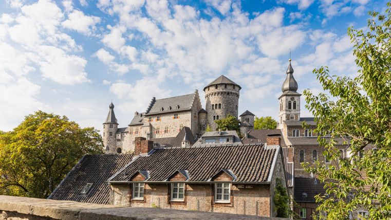 Een indrukwekkend kasteel met torens en kantelen, omringd door bomen. De lucht is blauw met enkele wolken.