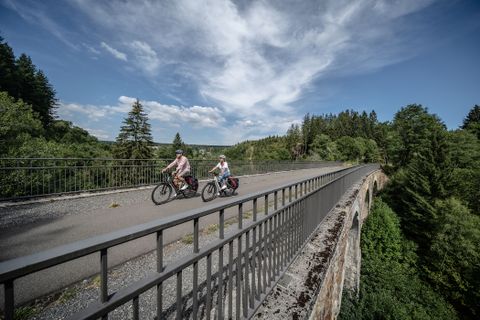 Zwei Radfahrer auf dem Vennbahn Viadukt Reichenstein, umgeben von Wald und blauem Himmel.