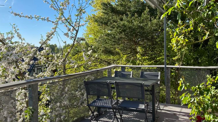 A cozy balcony with a table and chairs, surrounded by blooming trees. The sun is shining and the sky is clear.