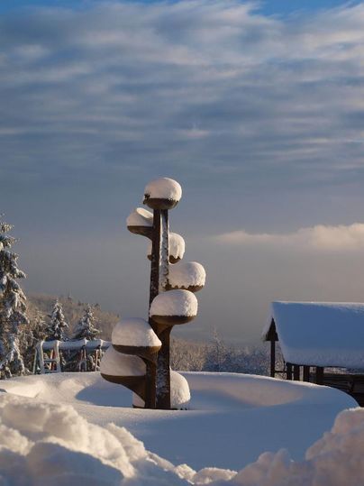 Eine verschneite Landschaft mit einem skulpturalen Holzstamm und sanften Hügeln im Hintergrund. Der Himmel ist teilweise bewölkt und hat eine sanfte, холодну Farbe.
