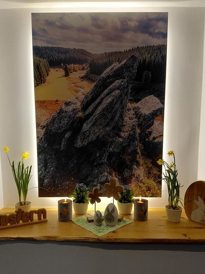 Ein schöner Holzregal mit einem Landschaftsbild im Hintergrund. Darauf stehen Blumen, Kerzen und österliche Dekorationen.