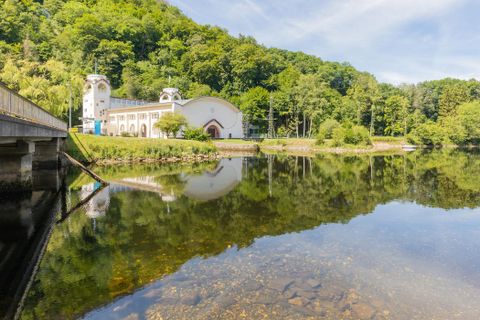 Ein ruhiger See spiegelt ein historisches Gebäude und grüne Bäume wider. Die Umgebung ist friedlich und idyllisch.