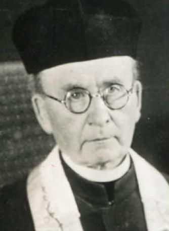 Un homme d'un certain âge, portant des lunettes et un couvre-chef noir. Il est vêtu de vêtements sombres et arbore une expression sérieuse.