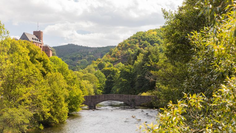 Eine malerische Landschaft mit einem Fluss und einer Bogenbrücke. Umgeben von grünen Bäumen und sanften Hügeln unter einem bewölkten Himmel.