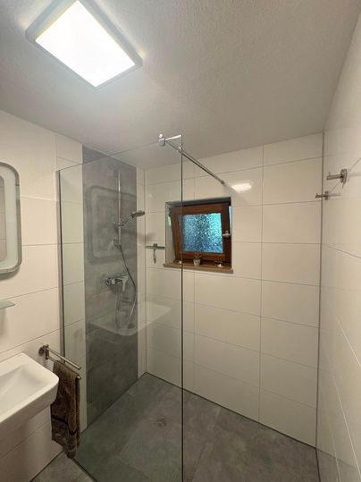 Une salle de bain moderne avec une douche en verre et une petite fenêtre. Les murs sont blancs et le sol est gris.