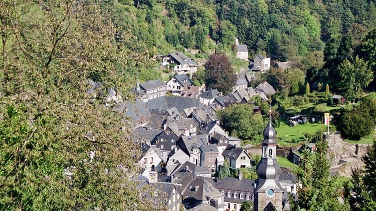 Ein malerisches Dorf in einem Tal, umgeben von grünen Wäldern. Die Dächer der Häuser sind dunkelgrau, und im Hintergrund ist eine Kirche zu sehen.