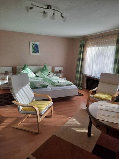 Ein gemütliches Schlafzimmer mit einem großen Bett und zwei Stühlen. Die Wände sind in sanften Farben gehalten und viel Tageslicht fällt durch die Fenster.