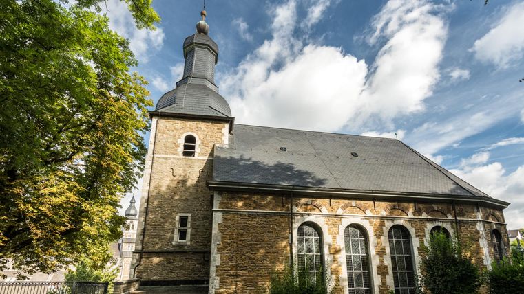 L'image montre une petite église avec un clocher caractéristique et de grandes fenêtres. Entourée d'espaces verts et d'arbres, elle dégage une atmosphère paisible.