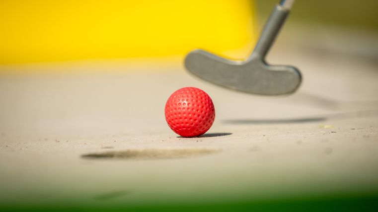 Een rode golfbal ligt op de grond. Een putter is op de voorgrond zichtbaar.
