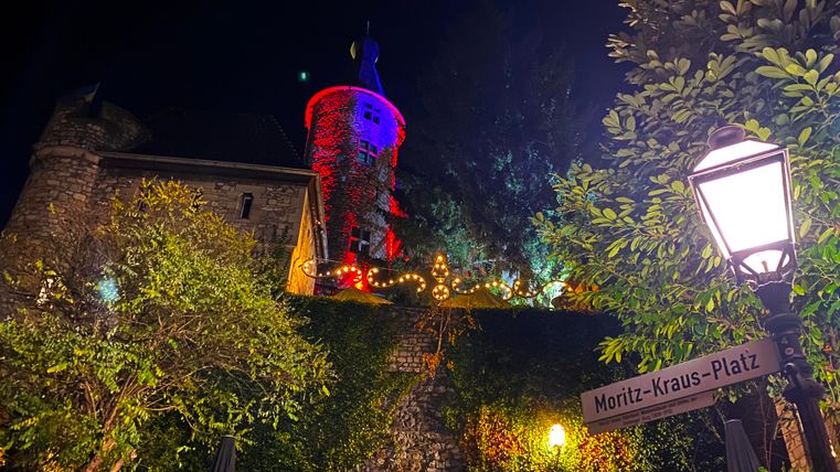 Eine Burg nachts mit bunten Lichtern verändert die Atmosphäre. Im Vordergrund steht eine Straßenschilder mit dem Namen Moritz-Kraus-Platz.