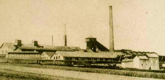 Eine alte Fabrikanlage mit hohen Schornsteinen und mehreren Gebäuden. Die Umgebung ist ländlich und weitläufig.