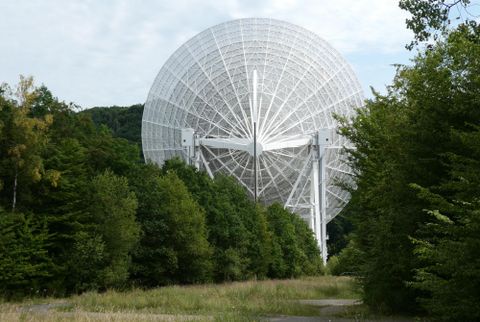 Eine große Satellitenschüssel steht zwischen Bäumen in einer grünen Landschaft. Der Himmel ist teilweise bewölkt.