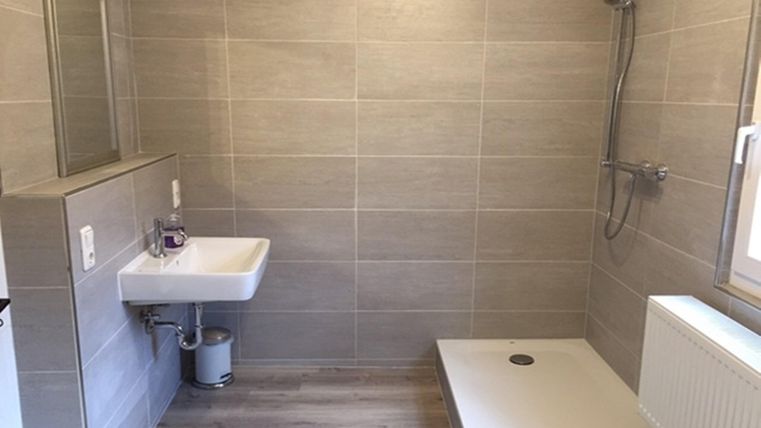 Une salle de bain moderne avec des carreaux gris et une douche de plain-pied. Le lavabo et un miroir sont également présents.