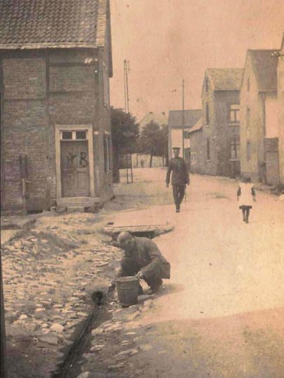 Une vue historique d'une rue pavée bordée de vieilles maisons. Un homme travaille par terre tandis qu'une personne et un enfant passent devant lui.