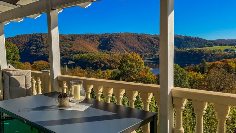 Eine gemütliche Terrasse mit einem Tisch und Sesseln bietet einen malerischen Blick auf die herbstliche Landschaft und den Fluss. Der klare blaue Himmel und die bunten Bäume sorgen für eine entspannte Atmosphäre.